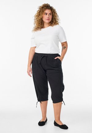 Kobieta o kręconych włosach, ubrana w białą koszulkę z krótkim rękawem, czarne spodnie 7/8 z troczkami i czarne płaskie buty, stojąca z jedną ręką w kieszeni.