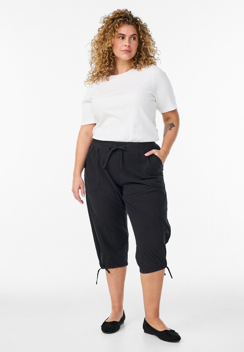 Žena s kučeravými vlasmi, ktorá má na sebe biele tričko s krátkym rukávom, čierne skrátené nohavice so sťahovacou šnúrkou a čierne baleríny, stojaca s jednou rukou v vrecku.