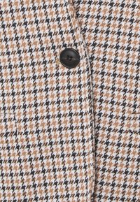 Zbliżenie na tkaninę w czarno-brązowo-białą kratę houndstooth i pojedynczy czarny guzik przyszyty do materiału.