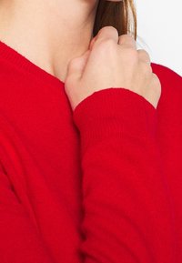 Roter Pullover aus weichem Strickstoff mit V-Ausschnitt und gerippten Bündchen. Nahaufnahme des Halsausschnitts und einer Hand, die den Stoff greift.