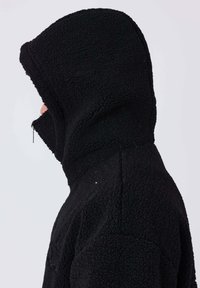 Personne de profil à gauche portant un sweat à capuche noir texturé avec la capuche couvrant entièrement la tête et un couvre-visage zippé.