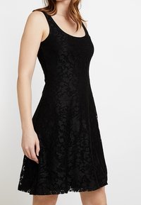 Robe en dentelle noire avec un corsage ajusté et une jupe évasée. Comprend un décolleté rond et des motifs floraux complexes. Texture lisse.