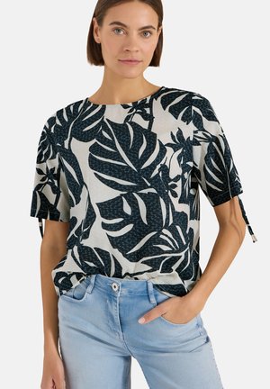 Femme portant un chemisier à manches courtes imprimé feuilles tropicales noir et blanc avec un jean bleu clair, debout, une main dans la poche.