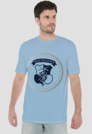 Man wearing light blue T-shirt with dark blue and gold Beauxbatons crest design, standing against plain light background.