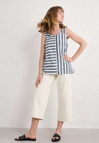 Niebiesko-biały w paski bezrękawnik o luźnym kroju, w zestawie z kremowymi szerokimi spodniami i czarnymi klapkami.