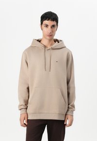 Sweat à capuche beige avec une poche kangourou, un cordon de serrage à la capuche, des poignets côtelés et un petit patch logo sur la poitrine. Fabriqué en tissu doux et texturé.