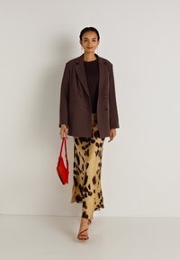 Bruine dubbelgebreide blazer over een aansluitend donker top, gecombineerd met een vloeiende beige rok met een zwart dierenprint en een rode handtas.