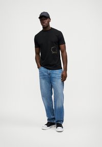 Μαύρο βαμβακερό t-shirt με ένα μικρό, κίτρινο γραφικό στο αριστερό στήθος, συνδυασμένο με ανοιχτό μπλε τζιν παντελόνι και μαύρα αθλητικά παπούτσια.