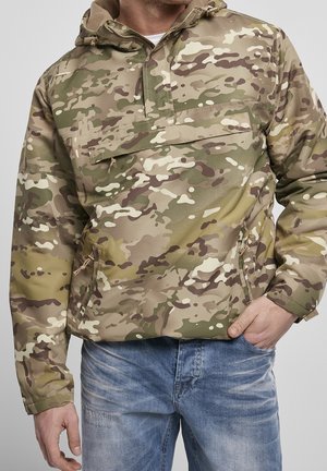 Man wearing camo-patterned hooded jacket with front pocket and blue jeans, hand in pocket, against plain background.