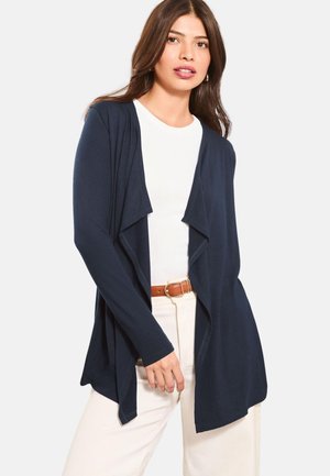 Nainen pukeutunut laivastonsiniseen avoimella edustalla olevaan neuletakkiin valkoisen paidan ja beigen housujen kanssa, ruskea vyö, seisoo vaalean yksivärisen taustan edessä.