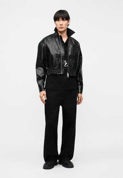 Νεαρός που φορά μαύρο κοντό μπουφάν από δερματίνη με υφή κροκοδείλου, μαύρη πόλο μπλούζα, φαρδιά μαύρη παντελόνια και μαύρα αθλητικά παπούτσια, στέκεται όρθιος.