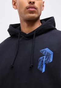 Εικόνα από κοντά ενός ατόμου που φοράει μαύρη κουκούλα με μπλε γραφικά βέλους και κείμενο «Βρες τη ροή σου, δες πού σε πάει η ελευθερία» στο στήθος.