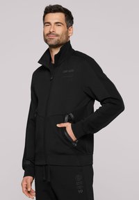 Zwarte zip-sweater met hoge kraag, voorzien van gaasaccenten en subtiele logo-details. Inclusief zijzakken en ribgebreide manchetten.