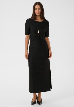 Femme portant une robe maxi noire texturée avec des manches aux coudes, une fente latérale, des talons noirs et un long collier avec un pendentif pompon.