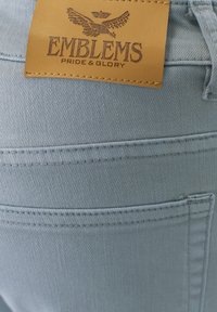 Tela de denim azul claro con una etiqueta de cuero que dice "EMBLEMS PRIDE & GLORY". Textura suave, costuras reforzadas y un bolsillo trasero visible.