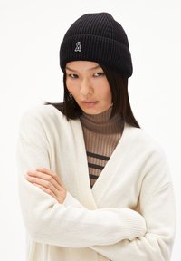 Schwarze gerippte Strickmütze mit einem kleinen gestickten Logo. Das Model trägt einen cremefarbenen Cardigan über einem braun gestreiften Rollkragenpullover.