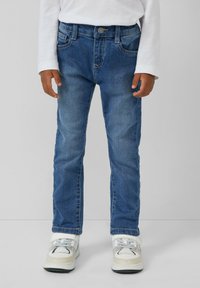 Dieťa oblečené v modrých denimových nohaviciach, bielej dlhý rukáv tričko a biele tenisky stojí na jednoduchom pozadí.