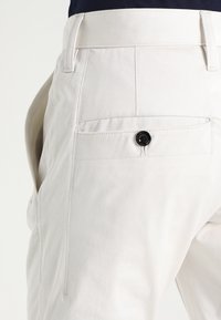 Vita bomullbyxor med en bakficka som stängs med en svart knapp, slät textur och enkel, skräddarsydd design.
