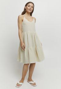 Helles Beige, knielanges Kleid mit einem gestuften Saum, V-Ausschnitt und dünnen Trägern aus strukturiertem Stoff. Kombiniert mit weißen Flip-Flops.