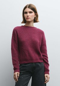 Suéter de punto color granate con textura acanalada, cuello redondo y mangas largas. Combinado con jeans de mezclilla oscura con detalles de tachuelas.