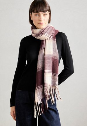 Femme aux cheveux courts bruns, portant un haut noir à manches longues et un jean foncé, avec une écharpe à carreaux bordeaux et crème enroulée autour du cou.