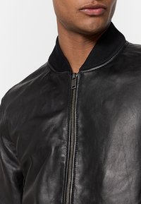 Giacca bomber in pelle nera con zip anteriore, colletto rotondo e dettagli a coste neri. La texture è liscia con una leggera lucentezza.