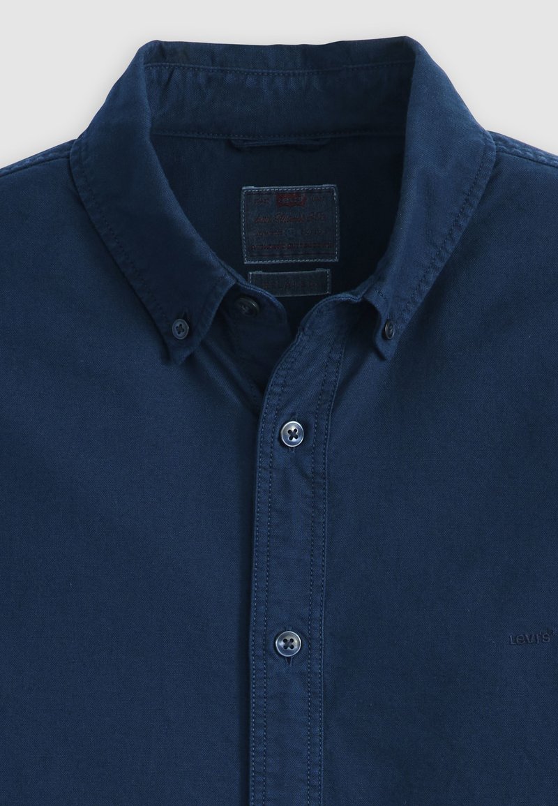 Camisa azul marino con botones y cuello abotonado, con etiqueta de marca visible dentro del cuello.