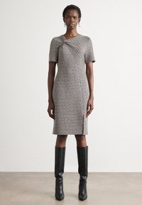 Robe jusqu'aux genoux avec un motif texturé noir et blanc, manches courtes et un détail de nœud sur le côté. Portée avec des bottes noires hautes.