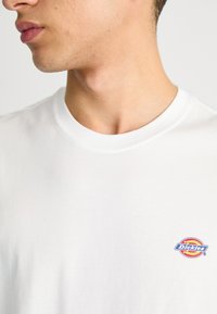 White cotton T-shirt with a crew neck and a small, colorful "Dickies" logo on the left chest area. Soft texture and simple design.