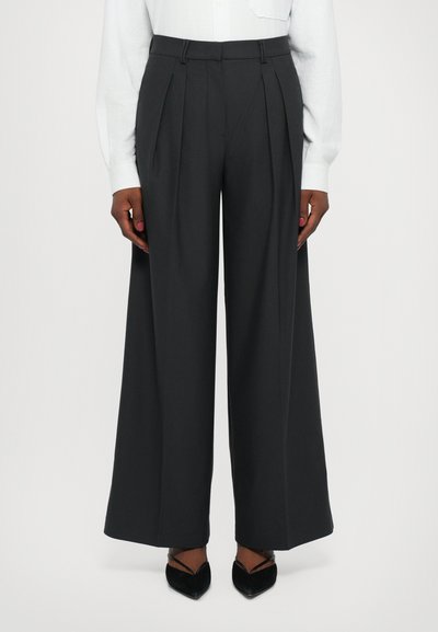Pantalones de pierna ancha en tejido gris oscuro, con pliegues frontales, una cinturilla ajustada y una textura suave. Combinados con zapatos negros de punta.