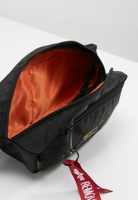 Black nylon travel pouch with an orange satin lining. Features dual zippered compartments and a red fabric tag with text.