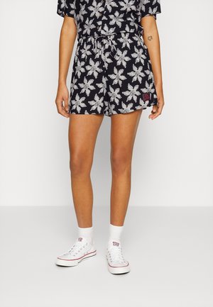 Person trägt schwarze Shorts mit weißen Sonnenmustern, weiße Socken und weiße Low-Top-Sneaker und steht vor einem einfarbigen Hintergrund.