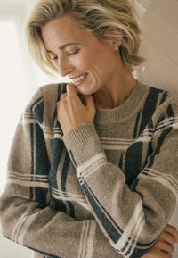 Beiger Wollpullover mit schwarz-weißem Karomuster, rundem Halsausschnitt und gerippten Bündchen. Das Modell berührt leicht mit einem sanften Lächeln ihr Kinn.