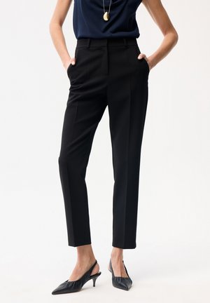 Osoba ubrana w czarne, dopasowane, sięgające kostek spodnie, granatowy, bezrękawnik, czarne, szpiczaste obcasy z paskiem z tyłu oraz złoty wisiorek na szyi.