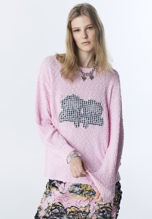 Joven con cabello largo y rubio, vistiendo un suéter rosa texturizado con una aplicación en blanco y negro, falda floral, collar con colgante de corazón plateado y pulseras.