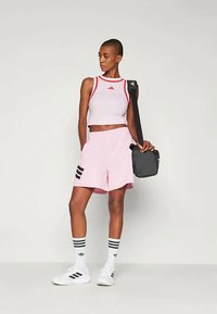 Ανοιχτό ροζ αθλητικές βερμούδες με μαύρες ρίγες, συνδυασμένες με λευκό crop top με κόκκινες λεπτομέρειες. Αξεσουάρ περιλαμβάνουν μια μαύρη τσάντα χιαστί.