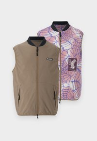 Reversible vest in brown fabric with side pockets; inner side features a pink-blue spider web pattern and brand logo patch.
