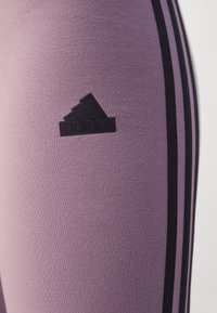 Leggings sportivi viola con un tessuto testurizzato, strisce laterali nere e un logo Adidas nero nella parte superiore della coscia.