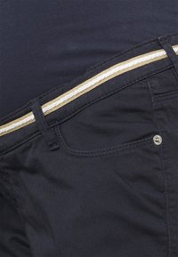 Gros plan sur un pantalon bleu marine foncé avec poche avant, passants de ceinture et ceinture style corde beige et blanche.