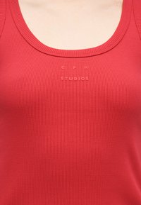 Κοντινό πλάνο ενός ατόμου που φοράει κόκκινη ριμπ τοπ χωρίς μανίκια με την ανάγλυφη επιγραφή "CPH STUDIOS" στην περιοχή του στήθους.