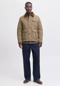 Hombre de pie con una chaqueta acolchada color beige, pantalones marino oscuros y zapatos marrones, contra un fondo claro.