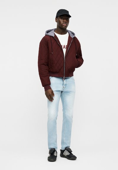 Jeune homme portant une veste matelassée bordeaux avec capuche grise, un t-shirt blanc à motif, un jean bleu clair, une casquette noire et des baskets noires à rayures blanches.