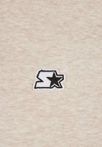 Patch brodé blanc et noir avec un « S » stylisé et une étoile sur un fond en tissu beige.