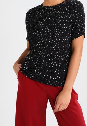 Woman wearing a black short-sleeve shirt with small white stars and loose red pants, one hand in pocket against a plain background.