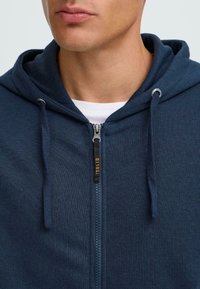 Mornarska zip-up dukserica od teksturiranog materijala, sa špagama na kapuci i patent-zatvaračem s oznakom brenda. Bijeli unutarnji sloj je vidljiv.