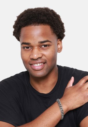 T-shirt noir, bracelet en chaîne argenté au poignet, teint moyen, cheveux noirs et bouclés, souriant, avec un fond clair.