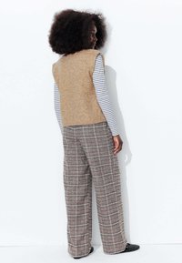 Gilet en laine beige sur une chemise à manches longues rayée blanche et noire, associé à un pantalon à carreaux larges dans des tons atténués. Chaussures noires à clous visibles.