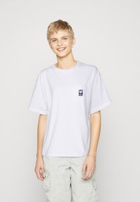 White cotton T-shirt with short sleeves, crew neck, and a left chest pocket featuring a blue graphic design. Worn with light gray cargo pants.