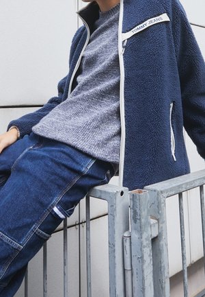 Personne appuyée sur une rambarde en métal portant une veste en polaire bleu marine Tommy Jeans, un pull en tricot gris et un jean bleu foncé avec des détails de poche.