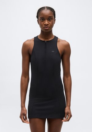 Frau mit geflochtenem Haar, die ein ärmelloses schwarzes Sportkleid mit Frontreißverschluss trägt und vor einem schlichten hellen Hintergrund steht.